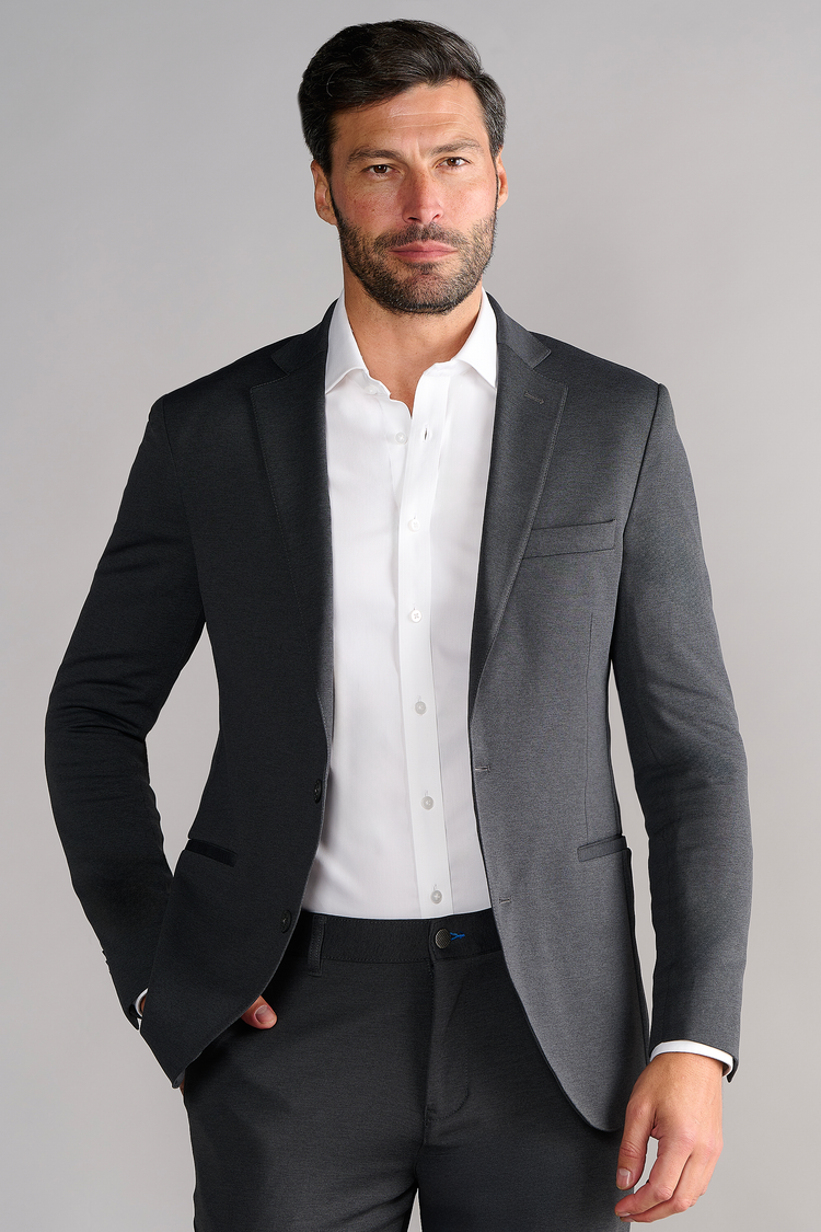 A man with dark hair and a beard wears the Twillory Circular Knit Blazer in Heather Grey over a white dress shirt. He stands facing forward with one hand in his pocket against a light gray background, showing a neutral expression. A man with dark hair and a beard wears the Twillory Circular Knit Blazer in Heather Grey over a white dress shirt. He stands facing forward with one hand in his pocket against a light gray background, showing a neutral expression.