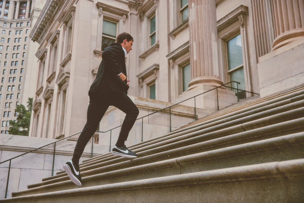 A man in a black suit and sneakers runs up the wide stone steps of a classical building.