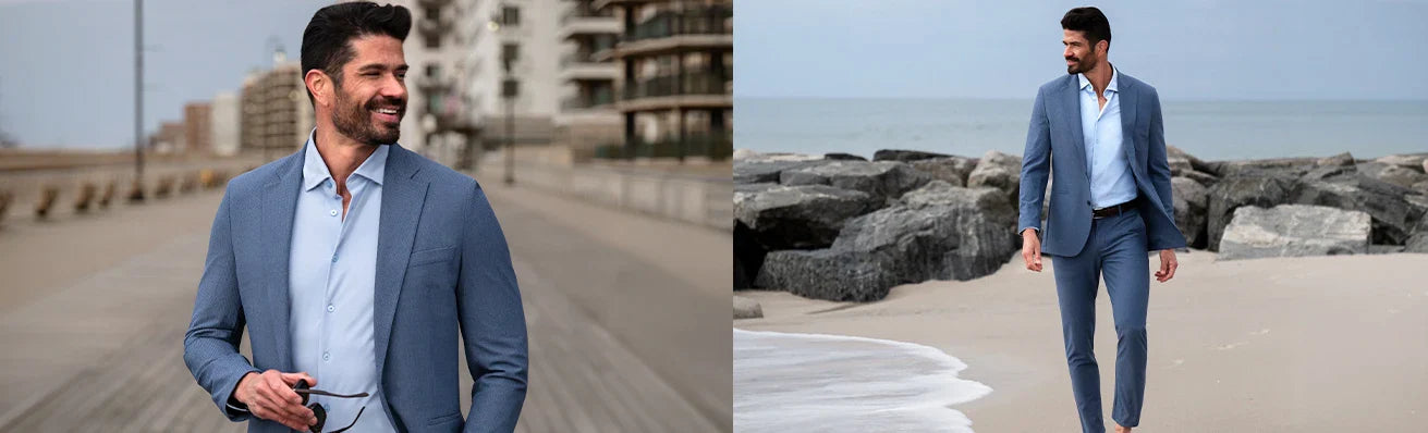 A man with dark hair and a beard wears a light blue suit and shirt, smiling on a boardwalk and walking barefoot on a rocky beach, holding sunglasses. The background shows buildings and the sea.