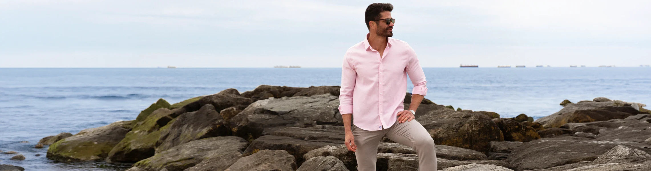 A man in sunglasses, a light pink shirt, and beige pants stands confidently on rocky shore, with the calm sea and distant ships under a cloudy sky in the background.