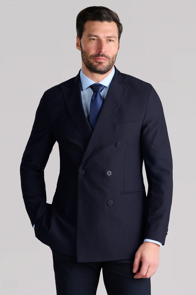 A man with short dark hair and a beard wears the Twillory AIR Double Breasted Blazer // Navy, paired with a light blue dress shirt and navy tie, standing confidently against a plain light grey background.