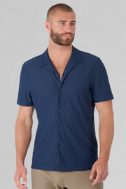 A man with short hair and a beard models the Twillory AIR Resort Shirt Solid in Navy, featuring short sleeves and cooling machine-washable fabric. He stands against a light gray background, looking to the side with a neutral expression. Only his upper body is visible.
