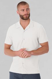 A man with a short beard and closely shaved hair wears the Twillory AIR Resort Shirt Solid in White, made from cooling AIR fabric. He stands against a light gray background, looking slightly left with hands clasped at his waist.