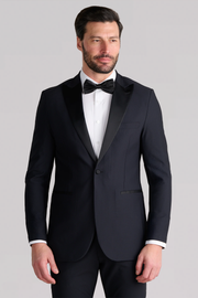 A man with short dark hair and a beard wears the Twillory AIR Tuxedo Blazer in black, featuring a breathable design and satin peaked lapel, paired with a white shirt and black bow tie. He stands against a light gray background, looking slightly to the side.