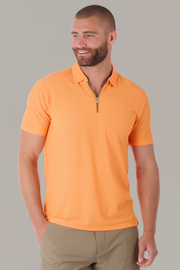 A man with a short beard and closely shaved hair smiles, looking left. He wears Twillory's AIR Zip Polo Solid in orange, featuring 360° Airflow Fabric, and light brown pants. His left hand is in his pocket against a light gray background.