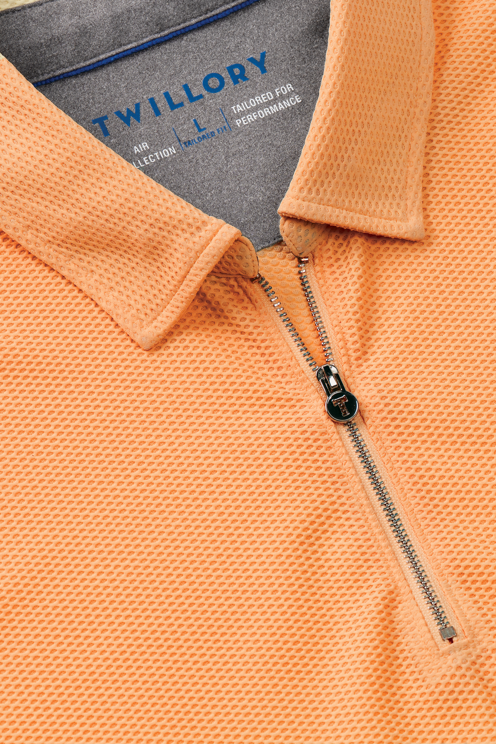 Close-up of the Twillory AIR Zip Polo Solid in orange, featuring a textured hyper-breathable fabric, silver quarter-zip with black pull tab, and an inside label detailing "Twillory AIR COLLECTION" and 360° Airflow Fabric.