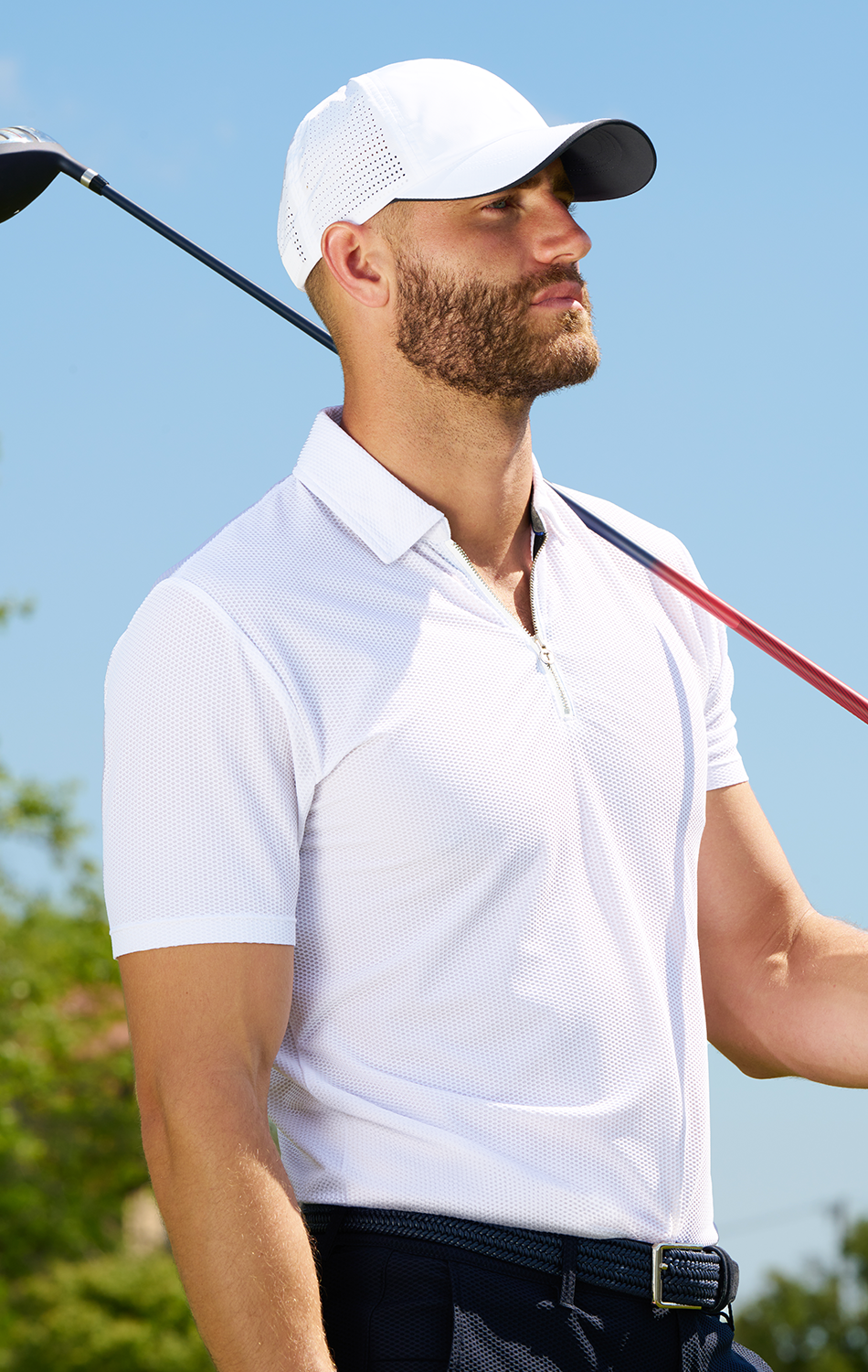 Man in a white polo shirt and baseball cap holding a golf club on a sunny day.