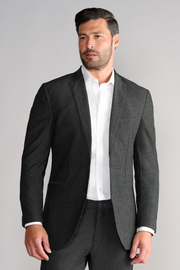 A man with short dark hair and a trimmed beard wears the Twillory AIR Blazer in Heather Grey over a white dress shirt, paired with matching pants. He faces slightly left against a plain light grey background, looking off-camera.