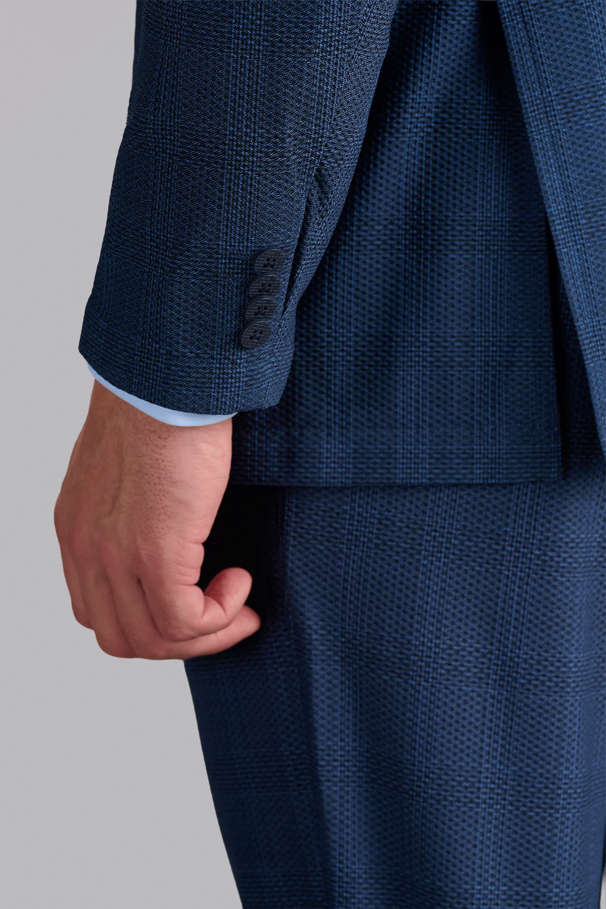 A close-up of a person’s left arm wearing the AIR Blazer // Blue Prince of Wales, featuring 360° Airflow Fabric with a subtle plaid pattern, three jacket sleeve buttons, and a light blue shirt cuff against a plain gray background.