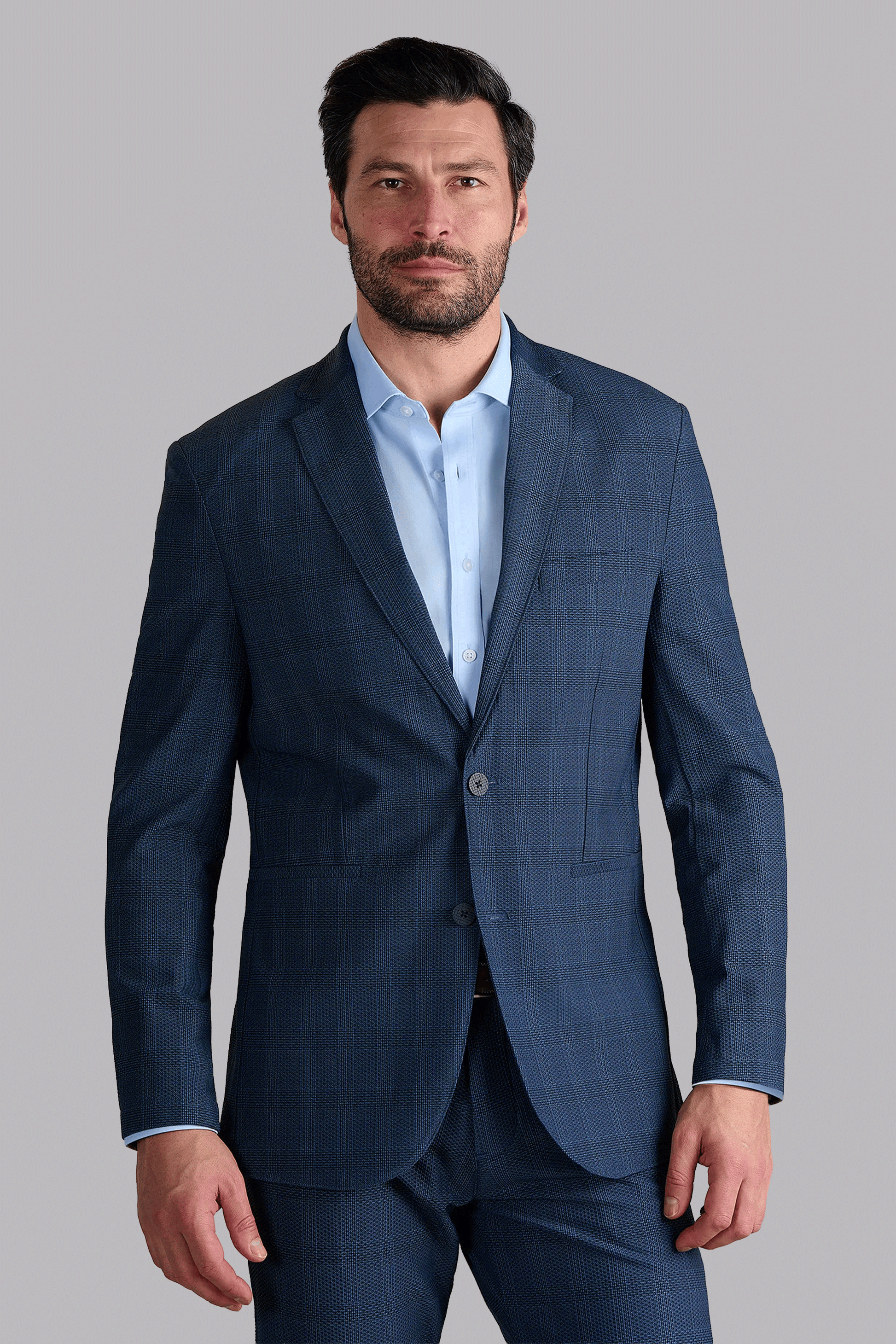 A man with short dark hair and a beard wears the AIR Blazer // Blue Prince of Wales, made with machine washable 360° Airflow Fabric. He stands against a light gray background, looking directly at the camera.