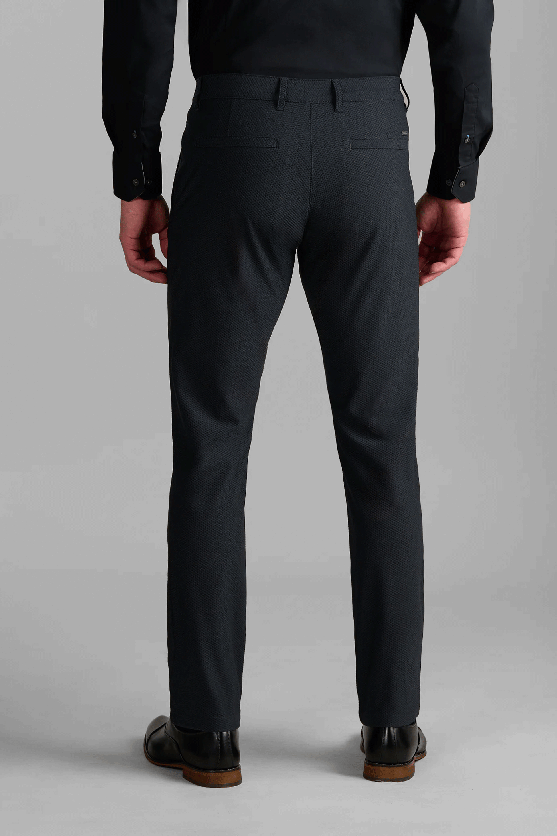 A person stands facing away, wearing Twillory AIR Pants in Heather Charcoal with two back welt pockets, paired with a black long-sleeve shirt and black dress shoes. The light gray background highlights their modern style.