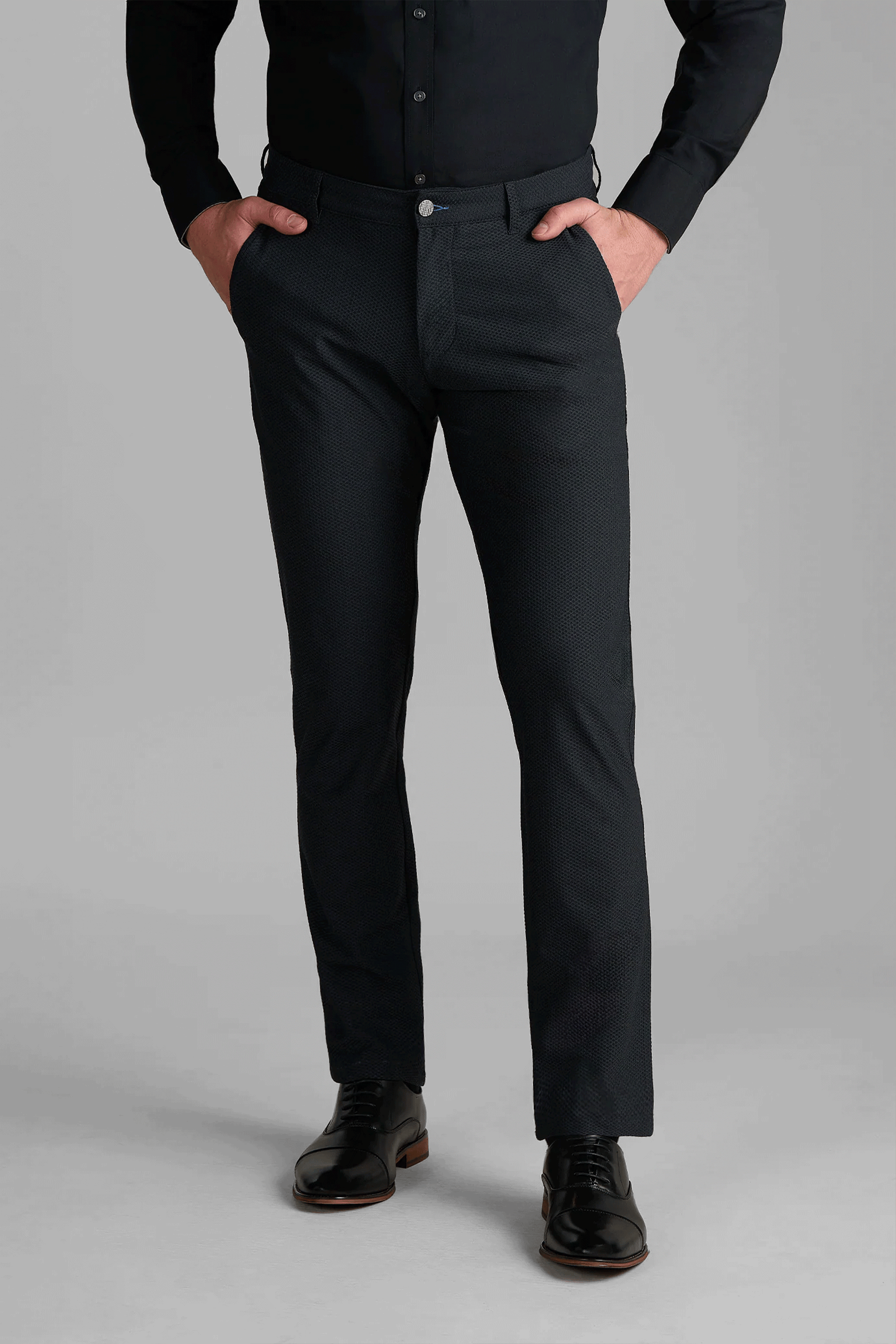 A person stands with hands in pockets, wearing Twillory AIR Pants in Heather Charcoal, a black button-up shirt, and shiny black dress shoes against a plain gray background. Only the body from shoulders down is visible.