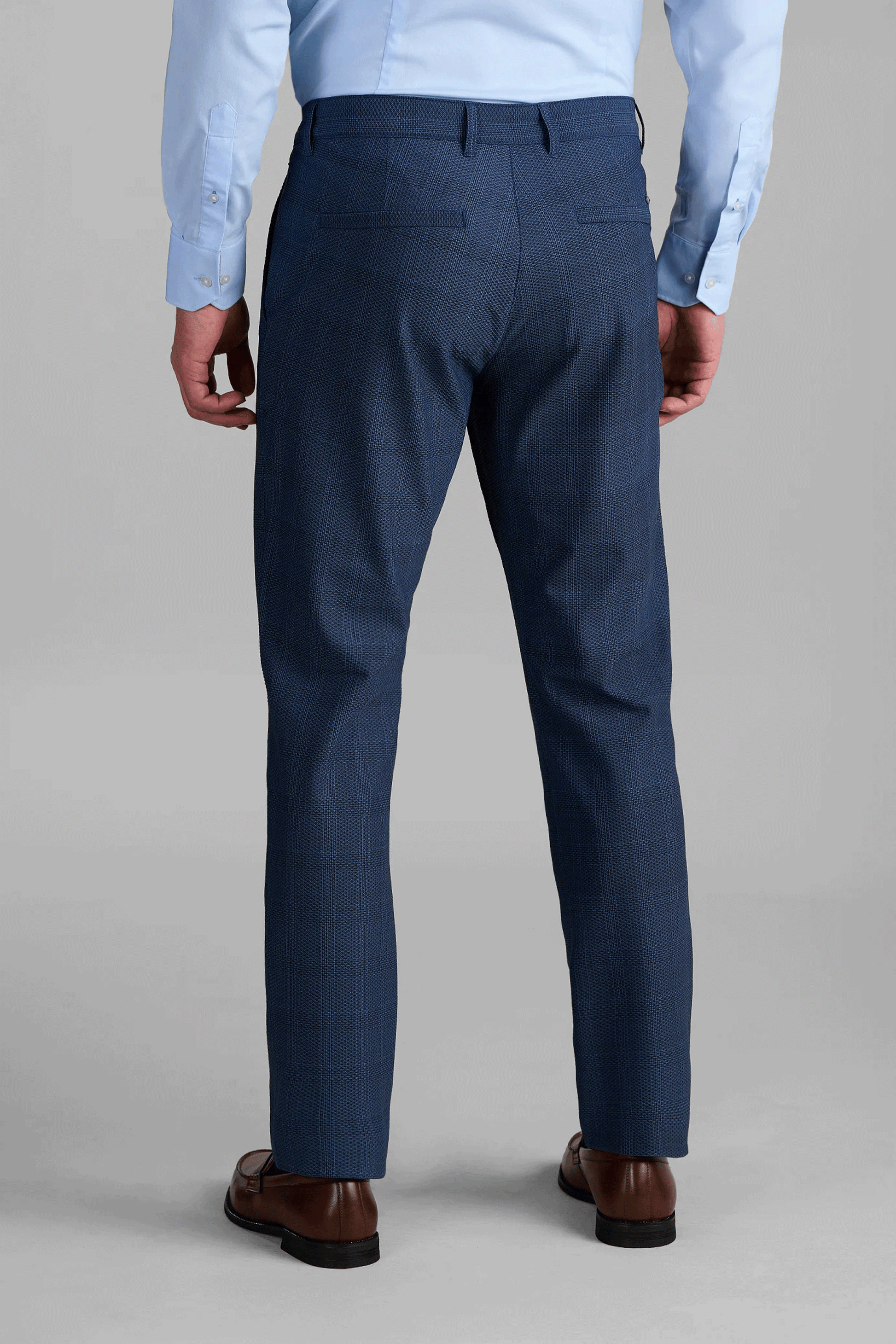 A man is shown from the back against a plain gray background, wearing a light blue dress shirt, brown dress shoes, and AIR Pants // Blue Prince of Wales.