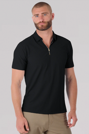 A man with a trimmed beard and short hair wears the Twillory AIR Zip Polo Solid in black, featuring 360° Airflow Fabric and a zippered collar, paired with tan pants. He stands against a light gray background, gazing to the side neutrally.