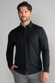 A man with short brown hair and a trimmed beard wears the Twillory Long Sleeve Button Down Polo Solid in black, paired with light gray pants. He stands against a plain light gray background, one hand in his pocket, looking to the side neutrally.