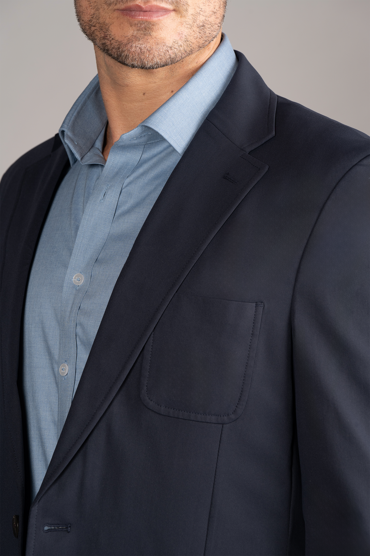 A man wears the Twillory Performance Patch Pocket Blazer in navy over a light blue dress shirt. The cropped image highlights the notched lapel and breast pocket, set against a plain light gray background.