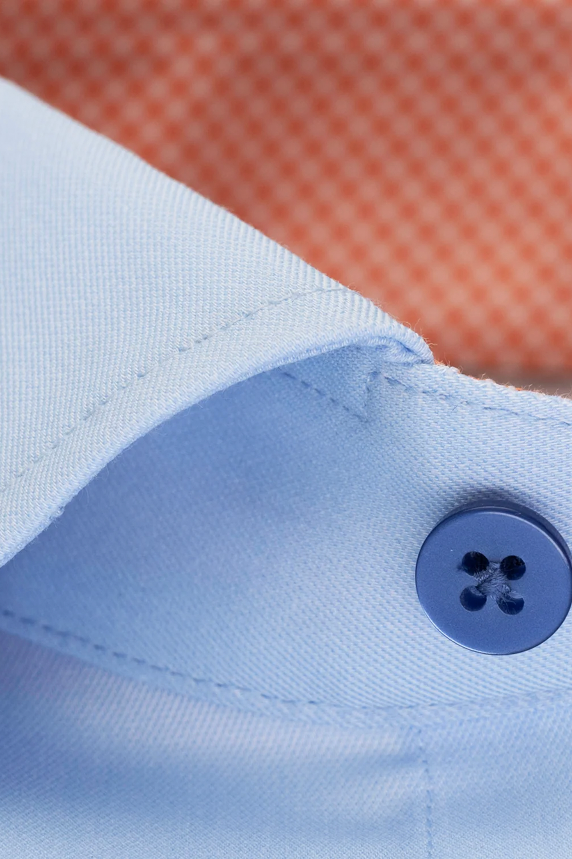 Close-up of the Twillory CLOSER // Performance Sunburst Contrast shirt’s light blue collar with a blue button. The blurred orange diamond-patterned background highlights the 4-way stretch comfort of this performance shirt.
