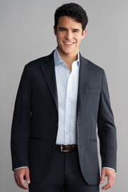 A young man with short, dark curly hair smiles against a gray background, wearing Twillory's Circular Knit Blazer in Black over a light blue dress shirt and dark trousers, accessorized with a brown belt.