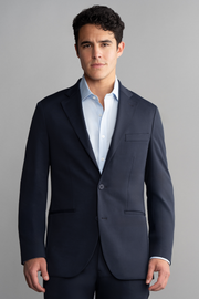 A man with short, dark curly hair faces the camera in a Twillory Circular Knit Blazer // Navy, featuring 4-way stretch, layered over a light blue dress shirt against a plain light gray background.