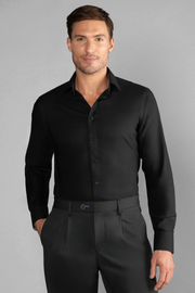 A man with short brown hair and light stubble, smiling softly with one hand in his pocket, wears Twillory’s The Event Shirt // Black—fitted, wrinkle-free officewear—tucked into matching black pants against a plain light gray background.