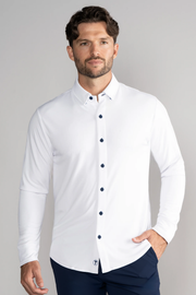 A man with short, wavy brown hair and a trimmed beard wears the Twillory Long Sleeve Button Down Polo Contrast in White Navy, dark blue pants, and stands against a light gray background with one hand in his pocket and a neutral expression.