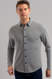 A man with light skin and short brown hair wears the Twillory Long Sleeve Button Down Polo Solid in Heather Light Grey, paired with dark jeans. He stands facing forward against a plain light grey background, his left hand slightly raised.