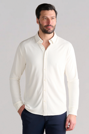 A man with short dark hair and a beard wears the Twillory Long Sleeve Button Down Polo Solid in Bone, styled with the top button undone and dark blue pants. He stands against a light gray background, one hand in his pocket, gazing to the side.