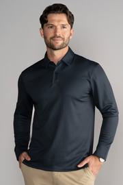 A man with wavy brown hair and a beard wears the Twillory Long Sleeve Polo Dot in Charcoal with light khaki pants. He stands against a plain gray background, smiling softly with his hands in his pockets and a watch on his left wrist.