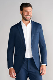 A man with short dark hair and a beard smiles against a light gray background, wearing Twillory's Performance Blazer in Navy Houndstooth over a white dress shirt, no tie, with his right hand relaxed at his side.
