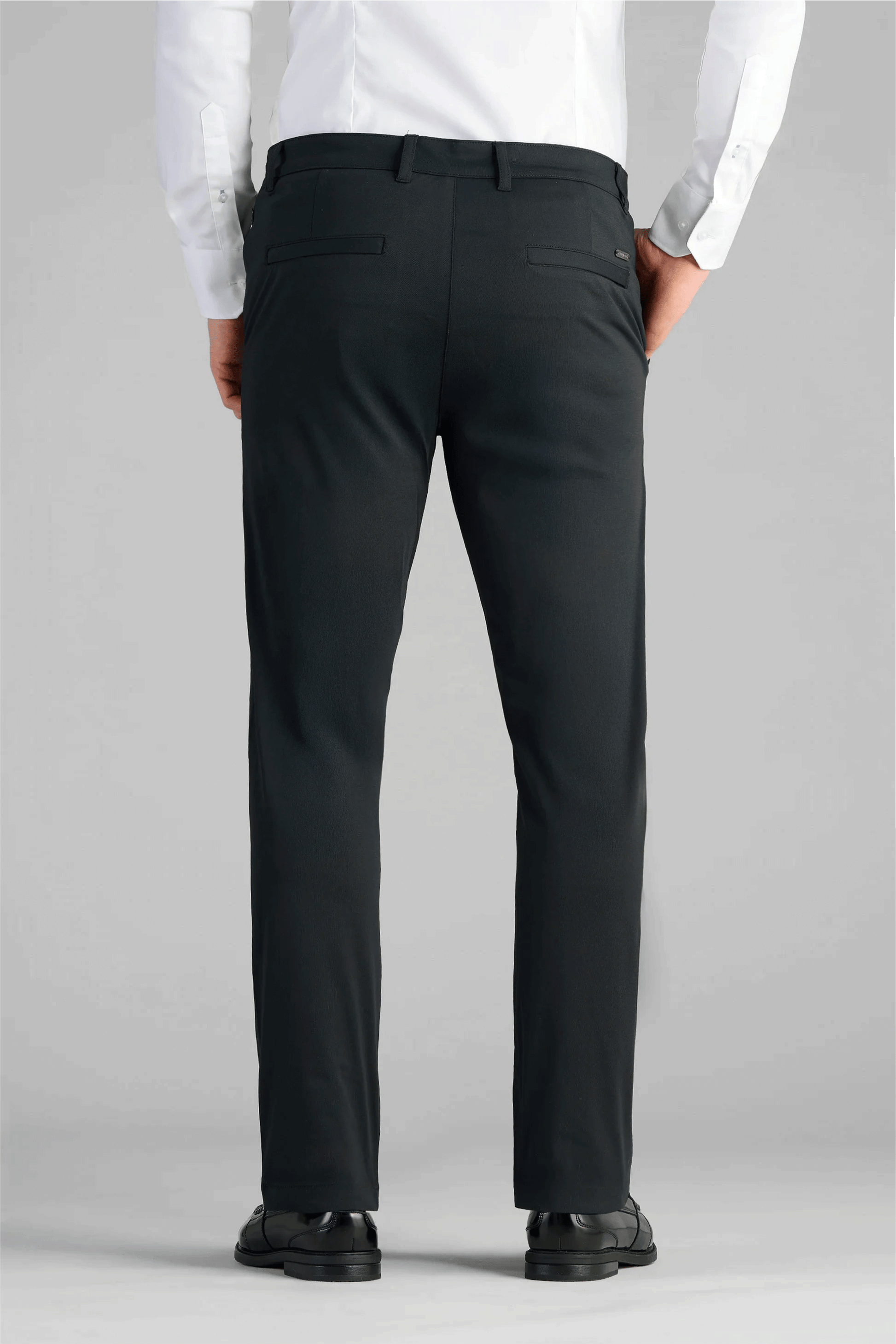 A person is seen from the back wearing a white dress shirt tucked into Twillory Performance Pants // True Black, featuring belt loops and welt back pockets, paired with black dress shoes. The background is plain light gray.