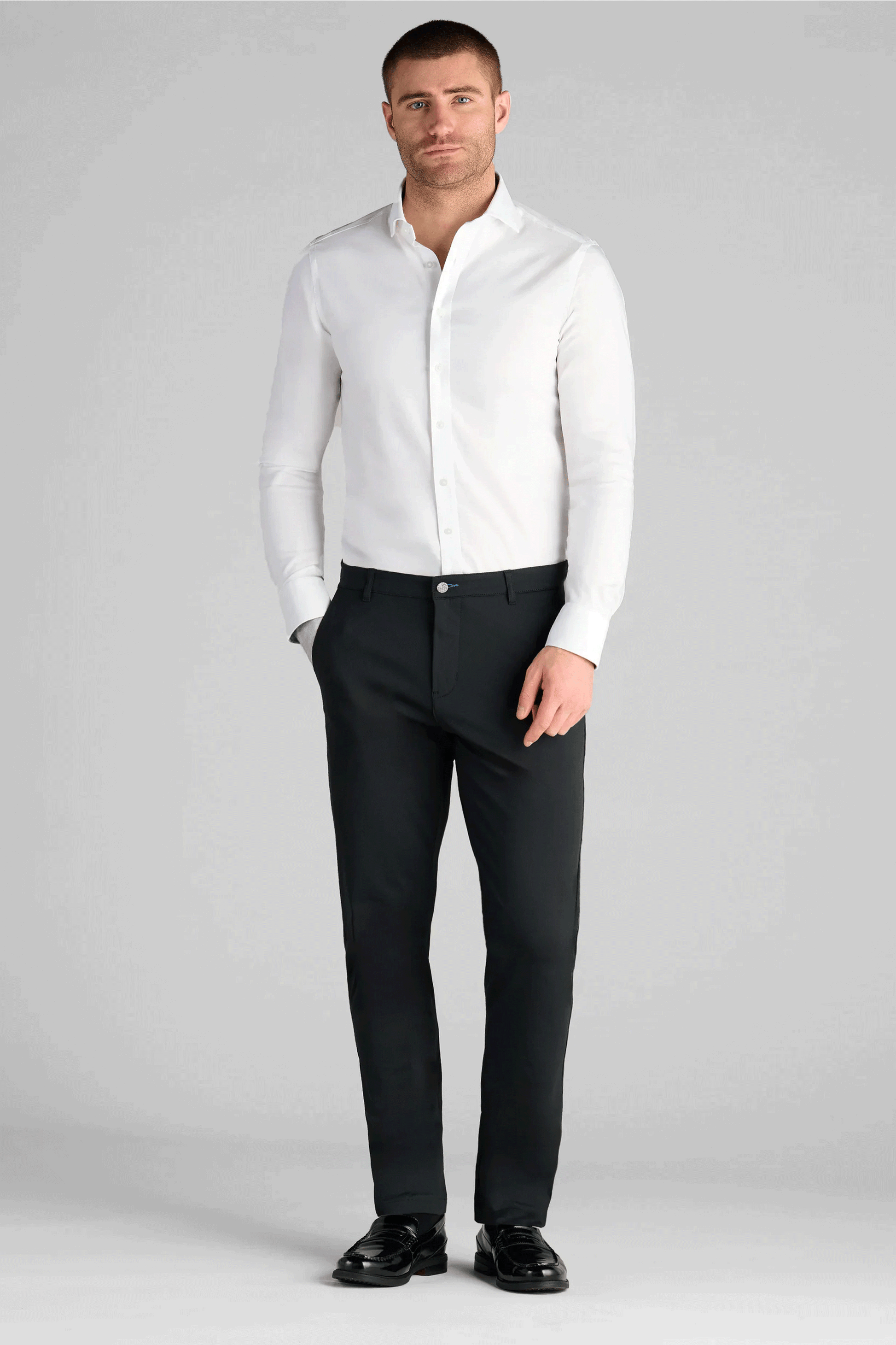 A man stands against a plain light gray background, wearing a fitted white shirt and Twillory Performance Pants // True Black with a shirt-gripping waistband. His left hand is in his pocket as he looks straight ahead with a neutral expression.