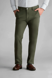 A person wears Twillory’s Performance Pants in Olive with a light blue button-up shirt and brown dress shoes, standing hands-in-pockets against a plain gray background. The image centers on the lower torso and legs; the head is not shown.