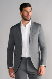 A man with short dark hair and a trimmed beard wears the Twillory Performance Blazer in Heather Light Grey over a white dress shirt, paired with matching trousers. He stands against a plain light gray background, looking to the side with a neutral expression.