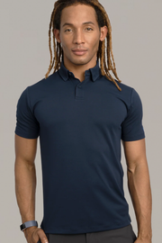 A person with long, light brown dreadlocks wears a Twillory Short Sleeve Polo Solid in navy and dark pants. They stand facing forward against a plain grey background, wearing a fitness tracker on their left wrist.