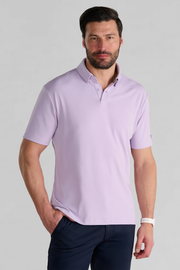 A man with short dark hair and a trimmed beard models the Twillory Short Sleeve Polo Solid in Lilac and dark navy pants. He stands against a light gray background, one hand in his pocket, wearing a white wristwatch.