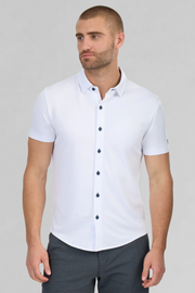 A man with short hair and a trimmed beard models the Twillory Short Sleeve Button Down Polo Contrast in White Navy, featuring dark buttons. He stands against a light gray background, gazing slightly left with a neutral expression, hands at his sides.