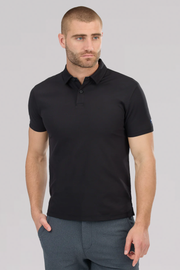 A man with short brown hair and a trimmed beard wears Twillory’s Slim Fit Pique Polo Solid in black and dark textured pants. He stands against a plain light grey background, one hand in his pocket, looking slightly to the side.