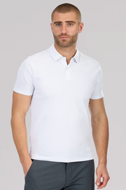 A man with short hair and a trimmed beard wears the Twillory Slim Fit Pique Polo Solid in white and dark pants, standing with his hands at his sides and looking neutrally at the camera against a light gray background.