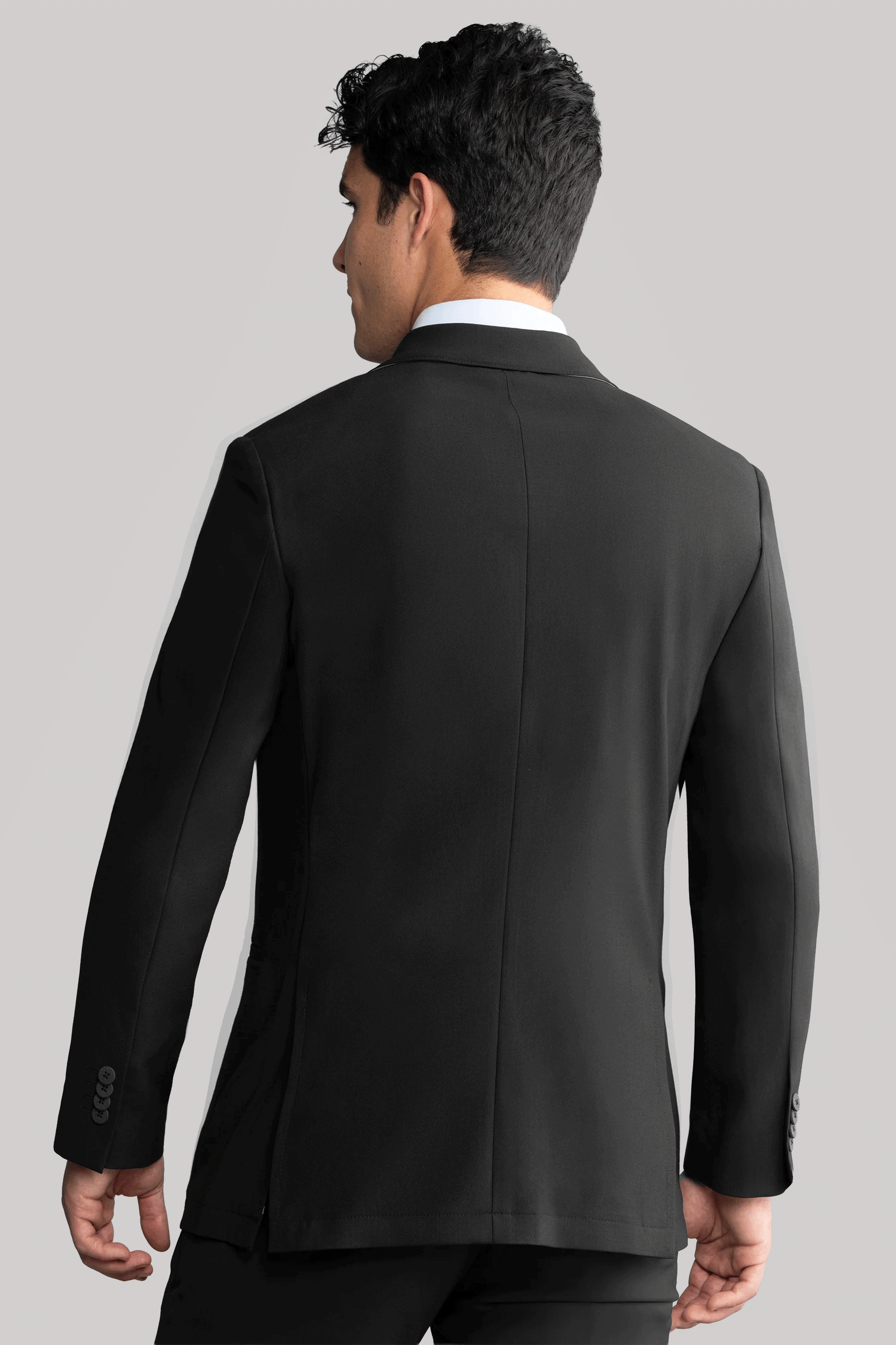 A man with short, dark hair wears the Twillory Hybrid Wool Blazer in black, featuring visible seams and cuff buttons, standing with his back to the camera against a plain light gray background.