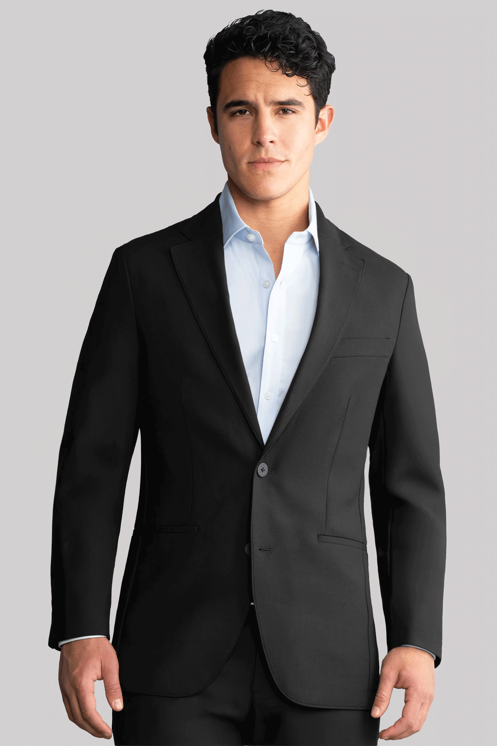 A young man with short, curly dark hair wears the Twillory Hybrid Wool Blazer in Black over a light blue dress shirt, standing forward with a neutral expression against a plain light gray background.