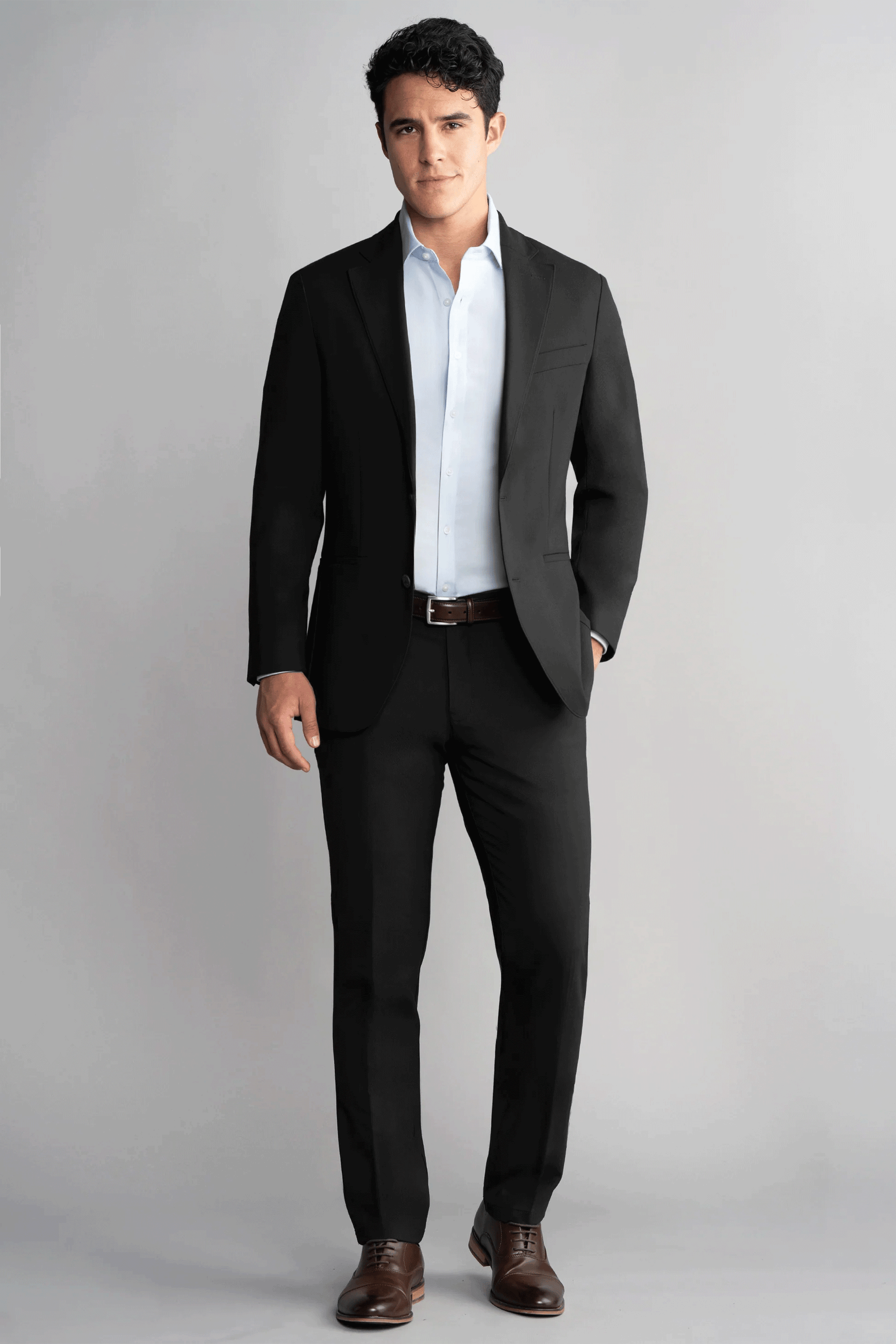 A man with short dark hair stands against a plain gray background, facing forward with a neutral expression. He wears Twillory’s Hybrid Wool Pants in black, paired with a light blue dress shirt and brown shoes, one hand in his pocket.