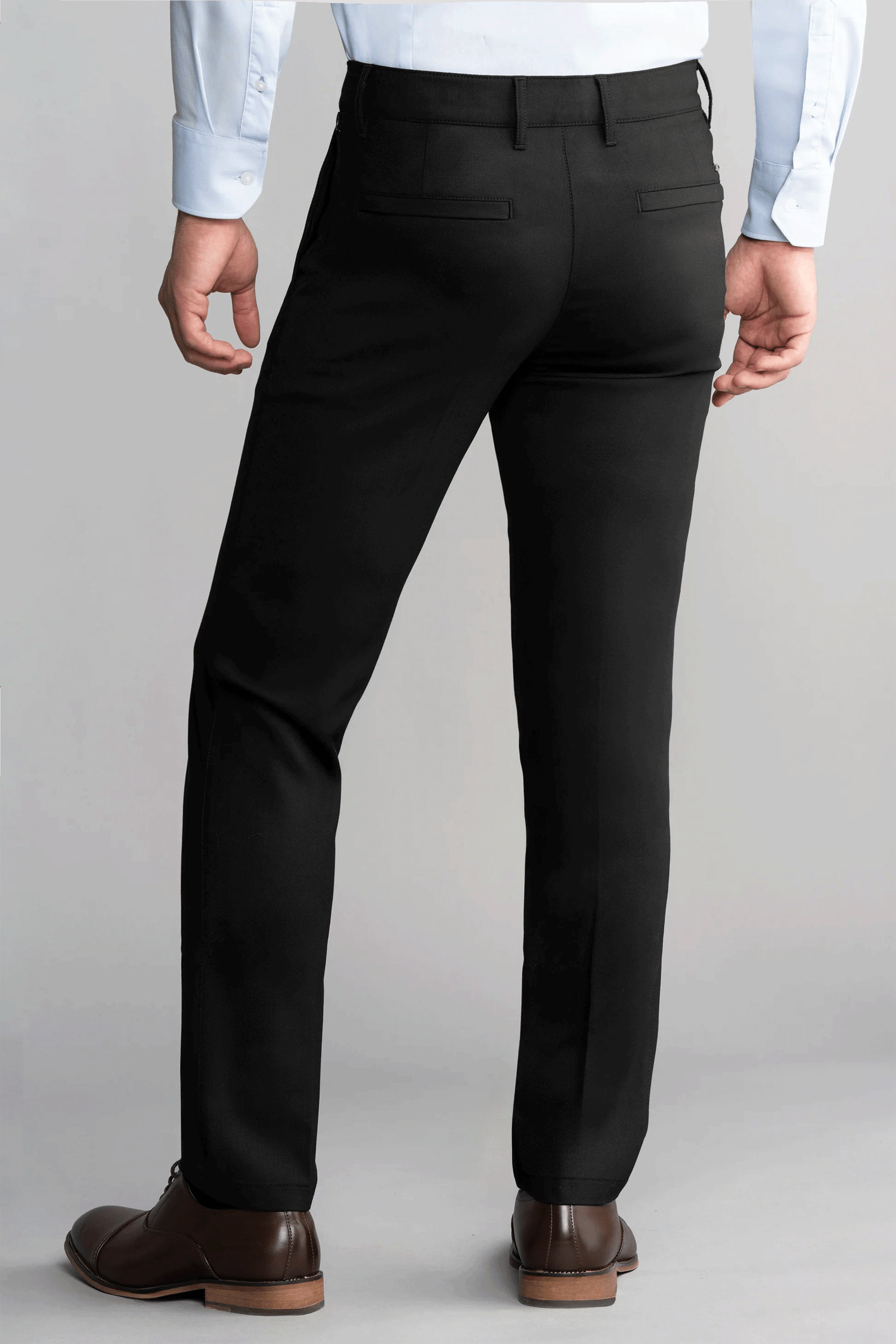 A man stands facing away in Twillory Hybrid Wool Pants // Black—slim-fit, 4-way stretch with two back welt pockets—paired with a light blue dress shirt and brown dress shoes, against a plain light gray background.