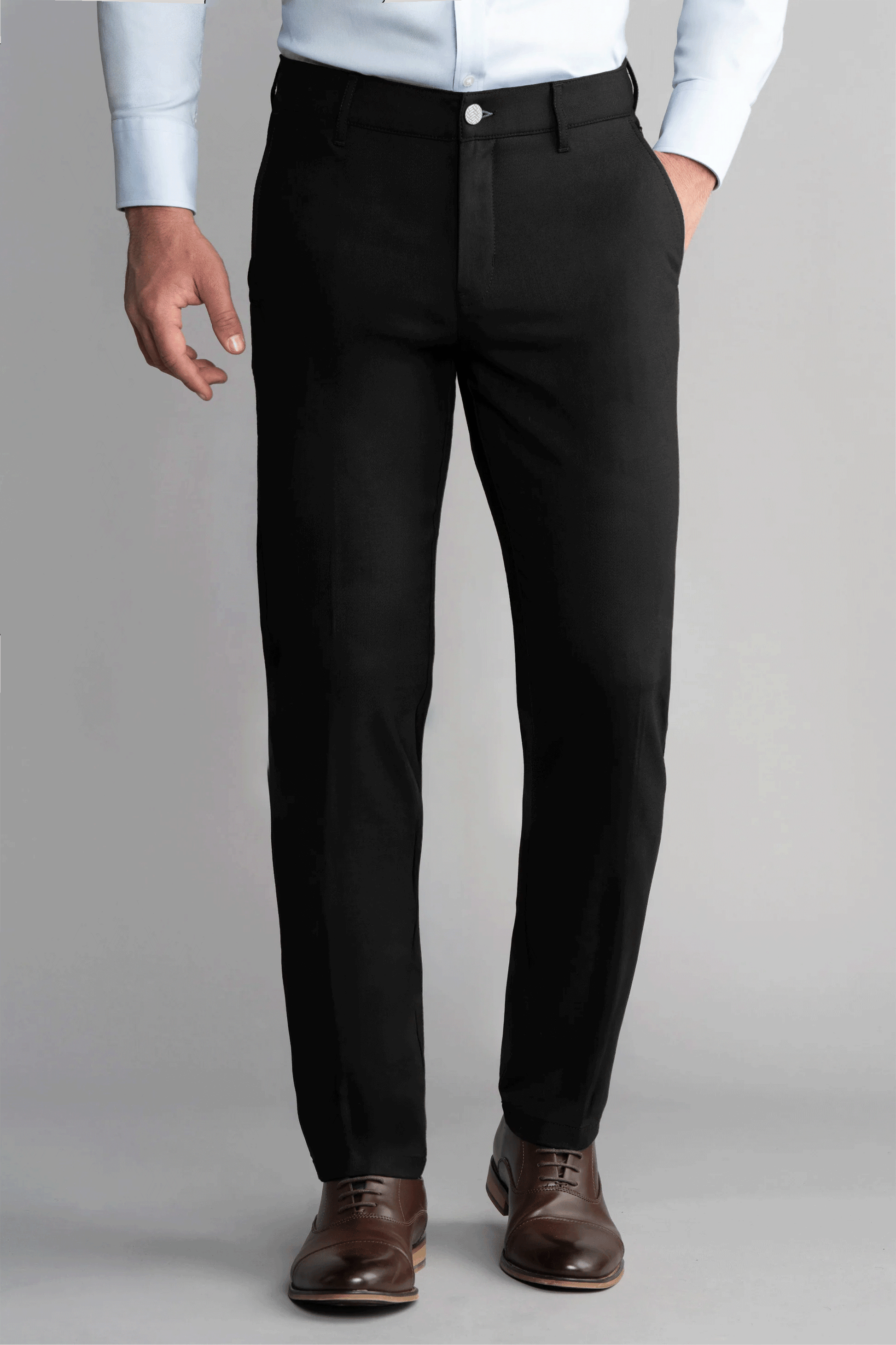 A man wears Twillory Hybrid Wool Pants in black, paired with a light blue dress shirt with rolled sleeves and brown leather dress shoes. He stands against a plain gray background, one hand in his pocket and the other relaxed by his side.