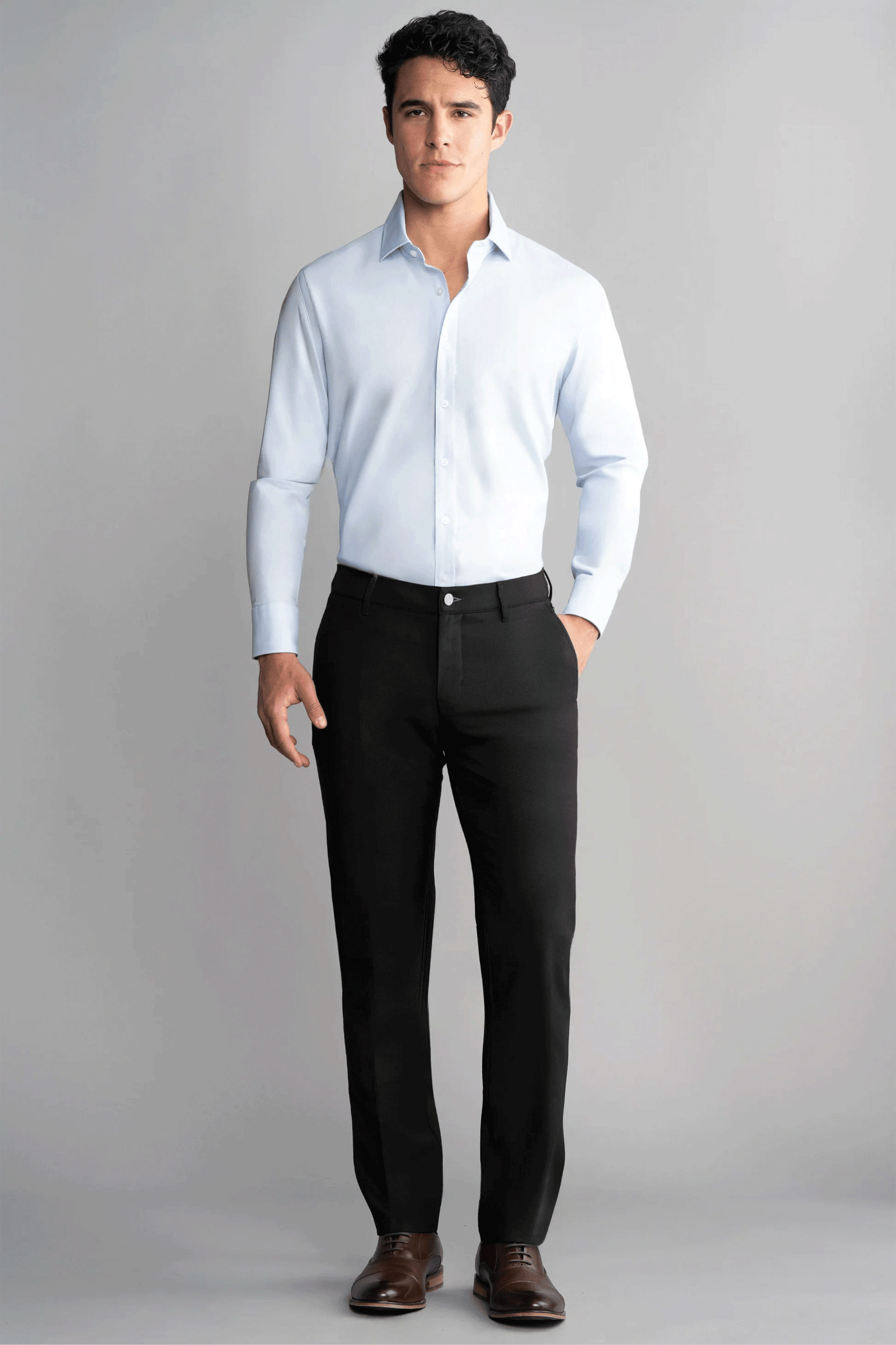 A man stands against a plain gray background wearing Twillory’s Hybrid Wool Pants in black. Crafted for comfort and style, he pairs them with a light blue dress shirt and brown shoes, one hand in his pocket as he looks directly at the camera.