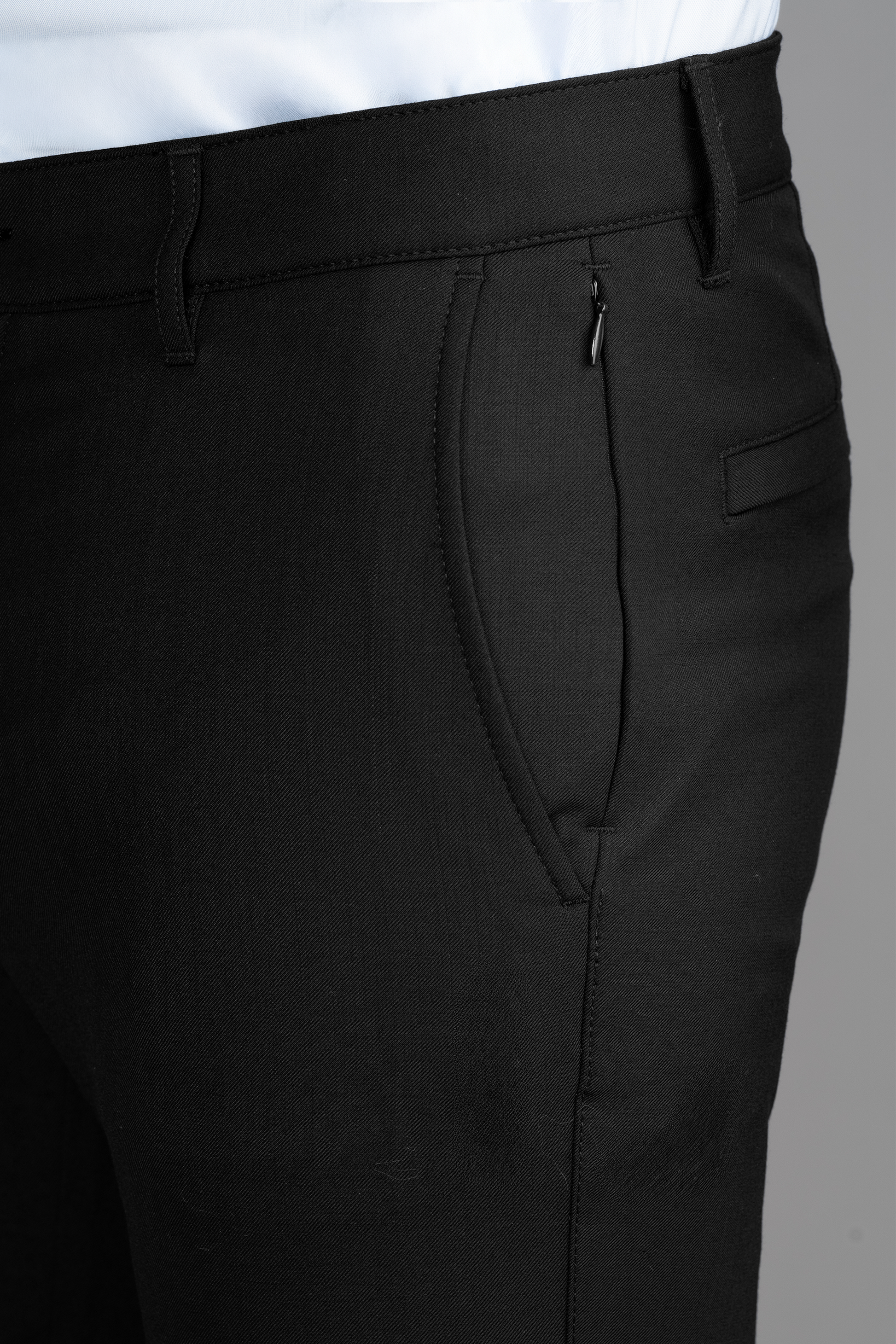 Close-up of Twillory Hybrid Wool Pants // Black, showing the waistband, belt loops, zippered side pocket, and a small horizontal slit pocket. A white shirt peeks above the waistband against a plain gray background.