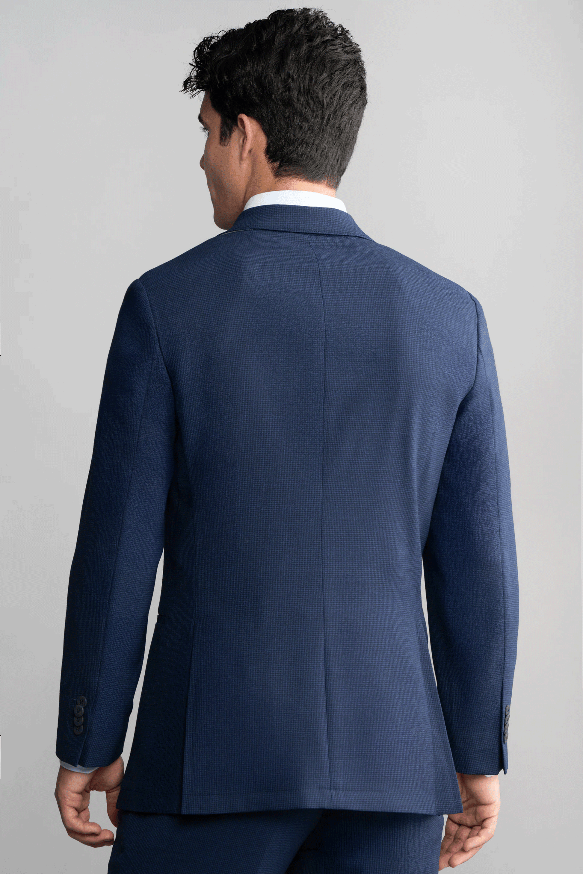 A man is seen from behind in a fitted Twillory Hybrid Wool Blazer // Navy Puppytooth and matching trousers. His wavy dark hair contrasts with the plain light gray background. The jacket offers 4-way stretch for comfort.