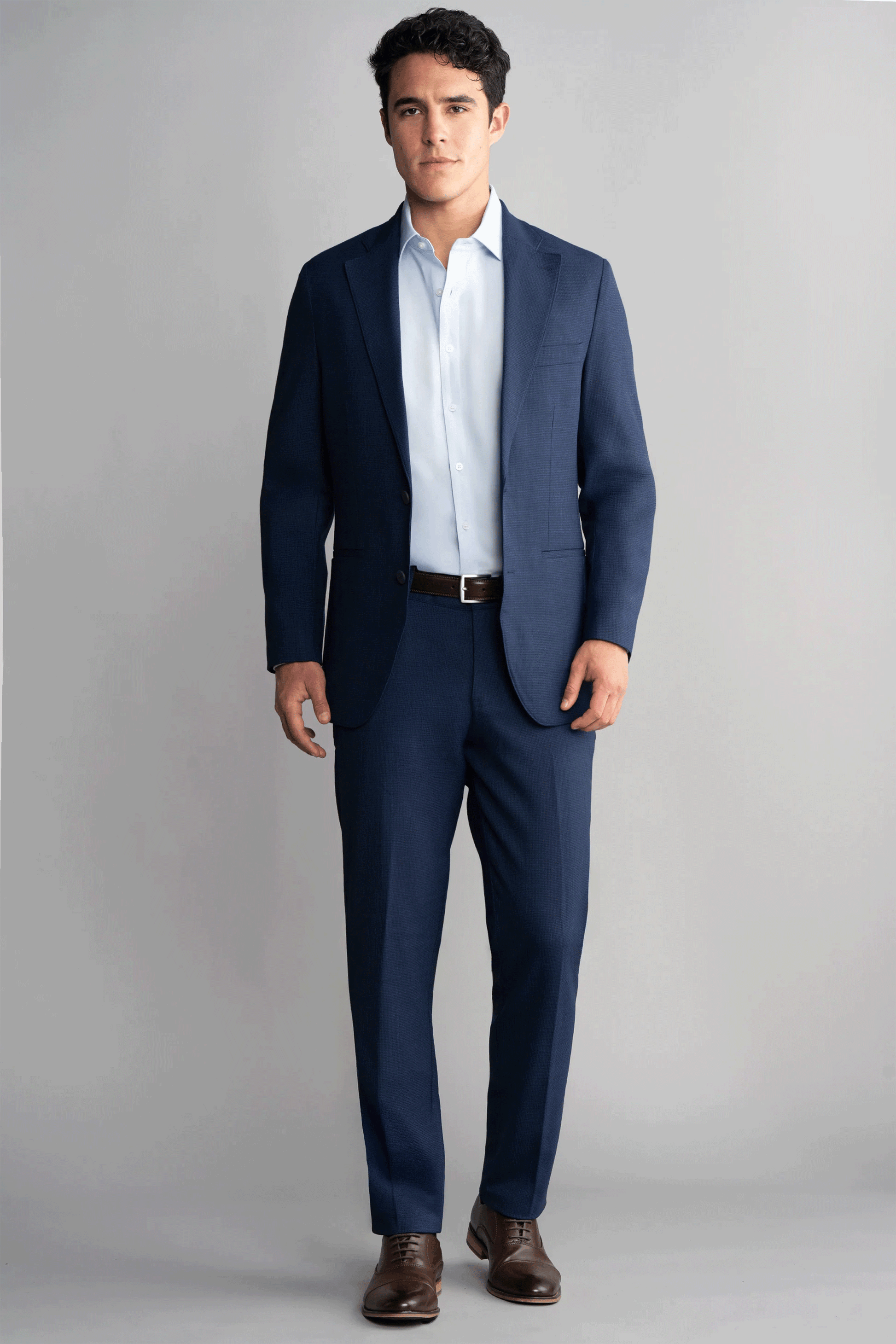 A man faces forward against a gray background, wearing Twillory's Hybrid Wool Blazer in Navy Puppytooth with a light blue shirt, brown belt, and brown shoes. His short wavy dark hair and neutral expression complete the look. The blazer is machine washable.