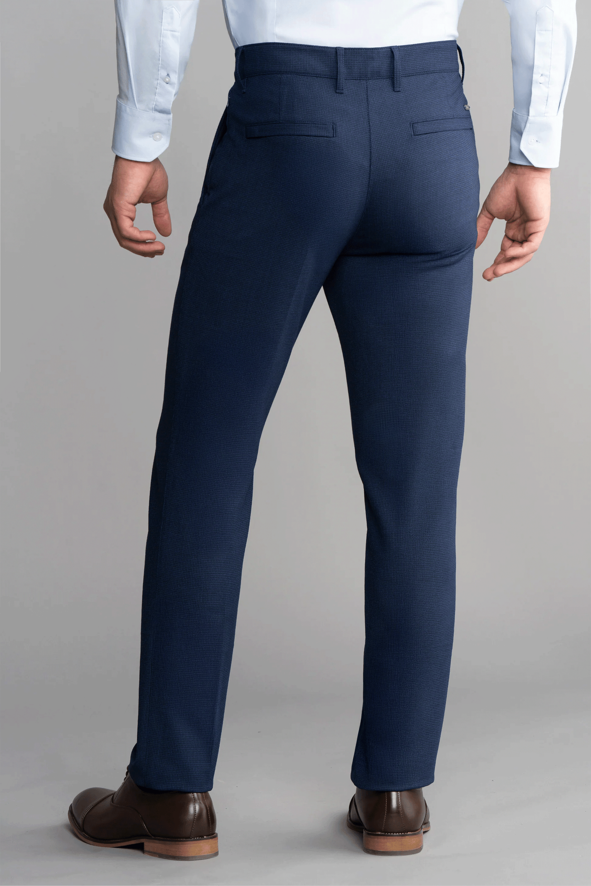 A person wearing Twillory Hybrid Wool Pants // Navy Puppytooth with 4-way stretch, a white dress shirt, and brown dress shoes stands facing away. The pants feature two buttoned back pockets against a plain gray background.