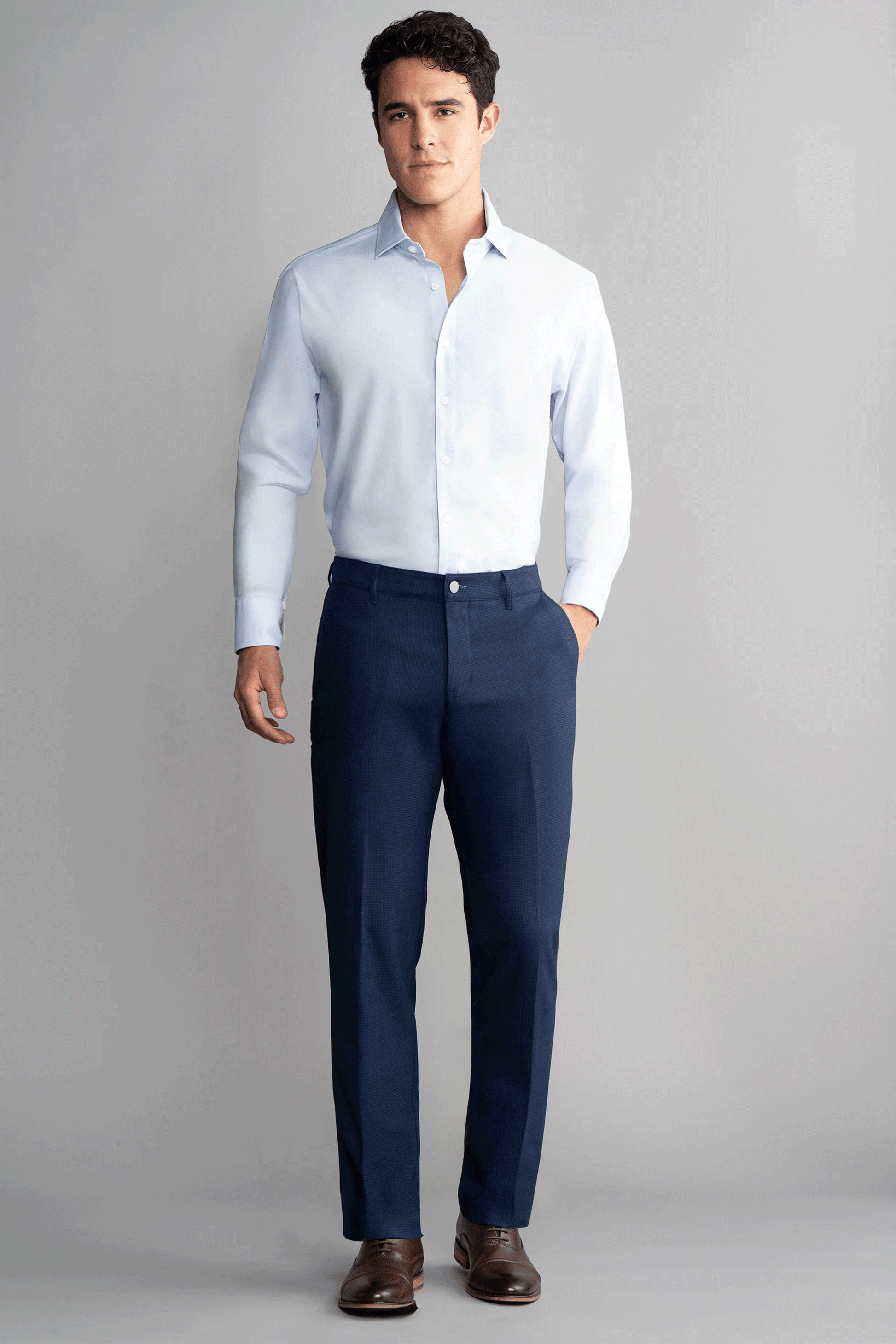 A man stands against a plain gray background wearing Twillory's Hybrid Wool Pants in Navy Puppytooth, paired with a light blue shirt and brown dress shoes. He has short dark hair, one hand in his pocket, and a neutral expression.