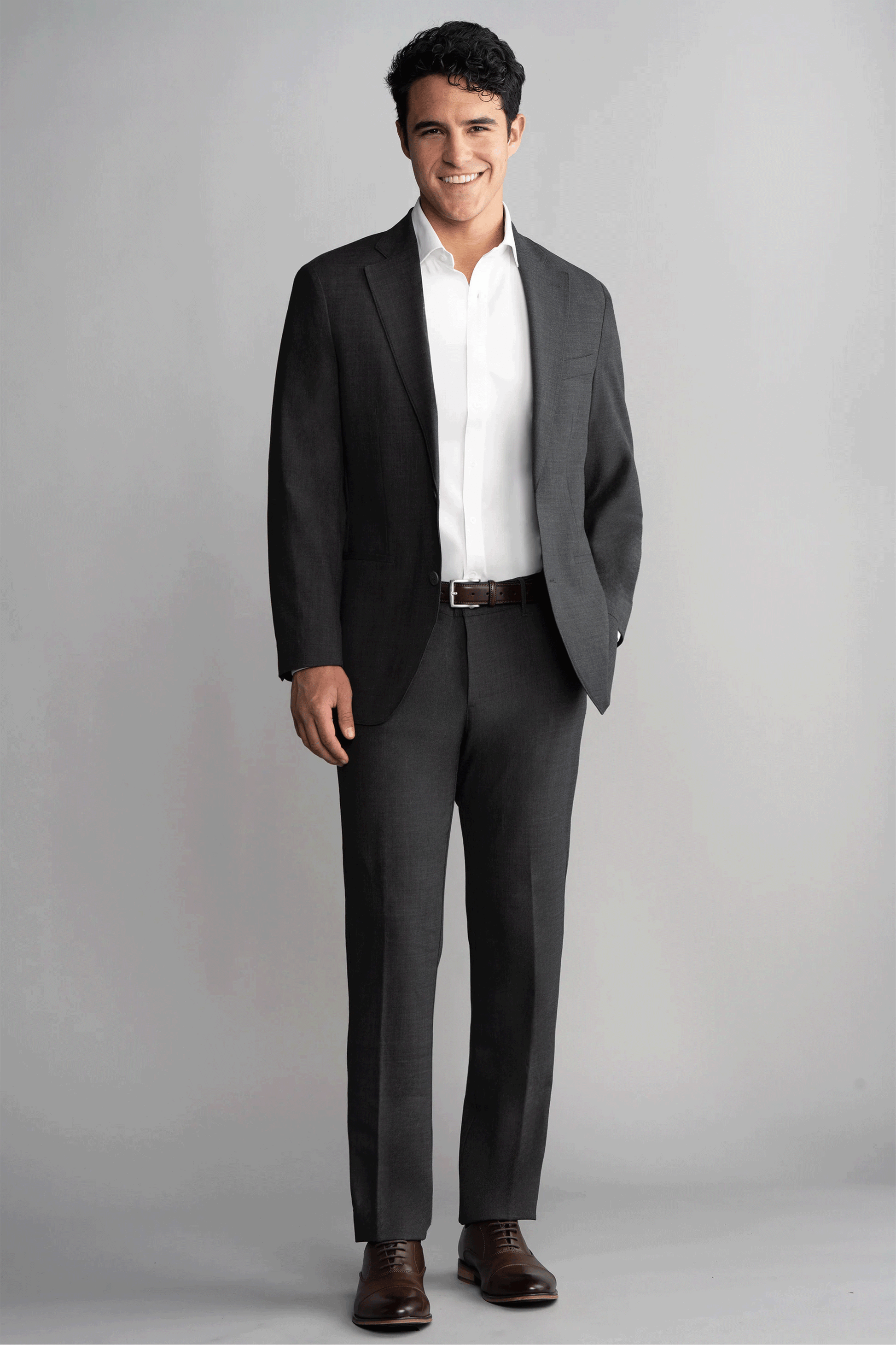 A man smiles against a light gray background, wearing Twillory's Hybrid Wool Pants in Grey Heather with a white dress shirt, brown belt, and brown shoes. His dark hair is neatly styled, and his hands rest casually at his sides.
